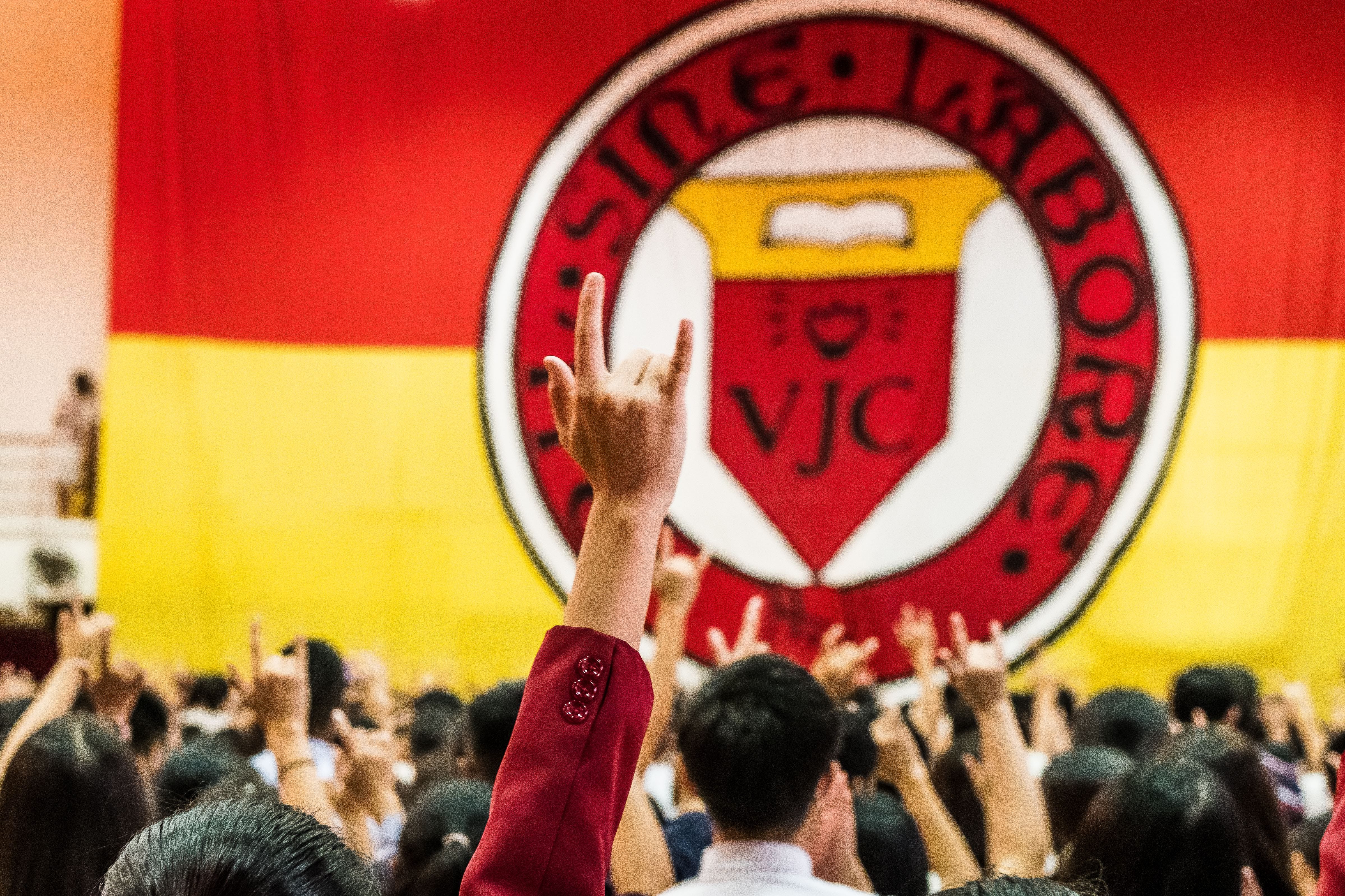 Crowd raising hands with rock signs before a large red and yellow VJC school crest banner.